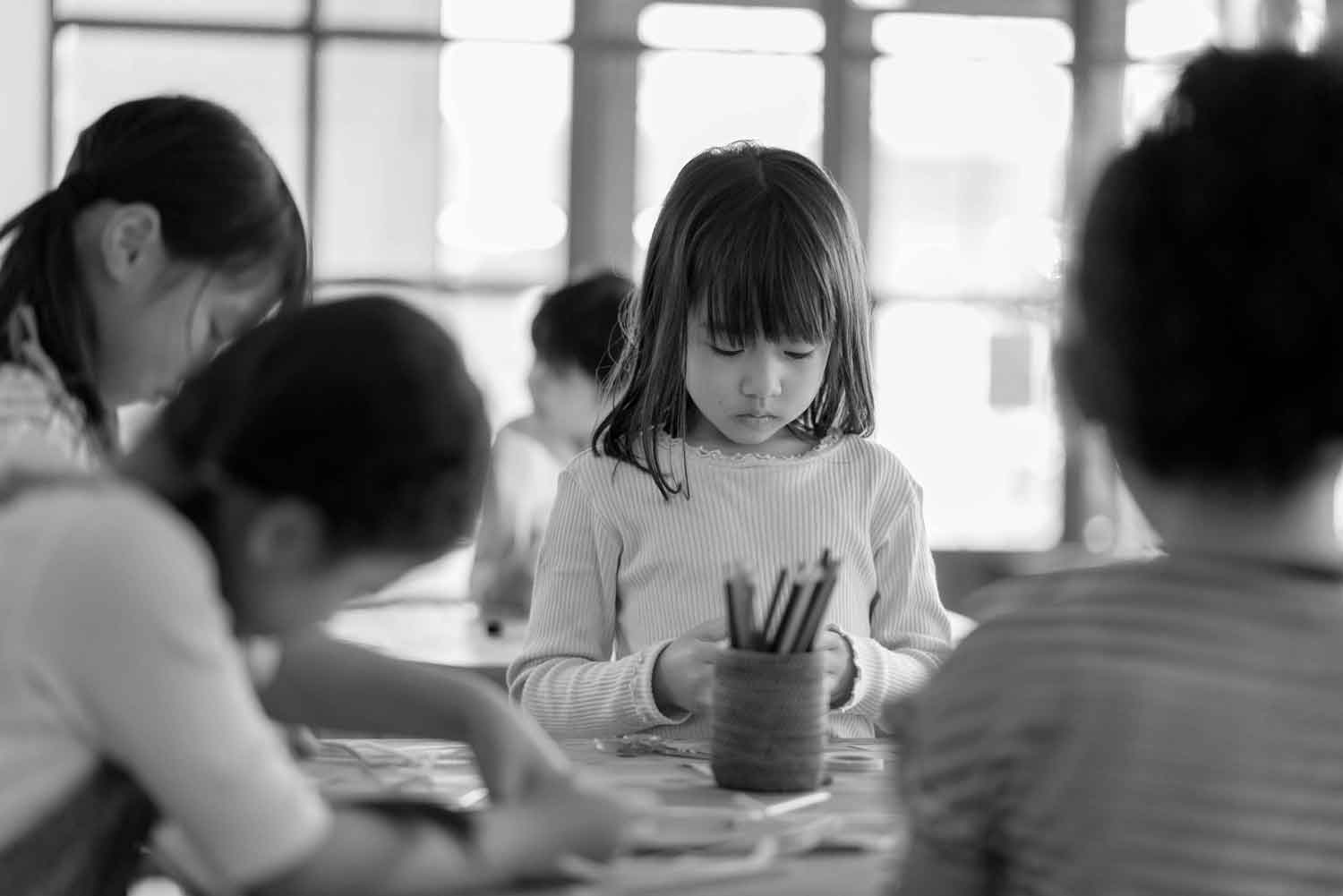 black and white image of young child drawing with pencils in front of her in a classroom setting