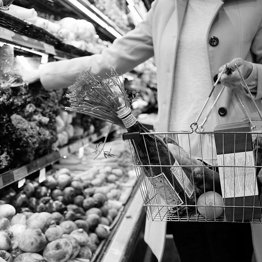 black and white image of woman holding wire shopping basket filled with fruit and a bottle of water