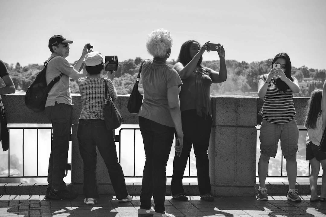 black and white image of group of people taking photos in front of beach and hills in background