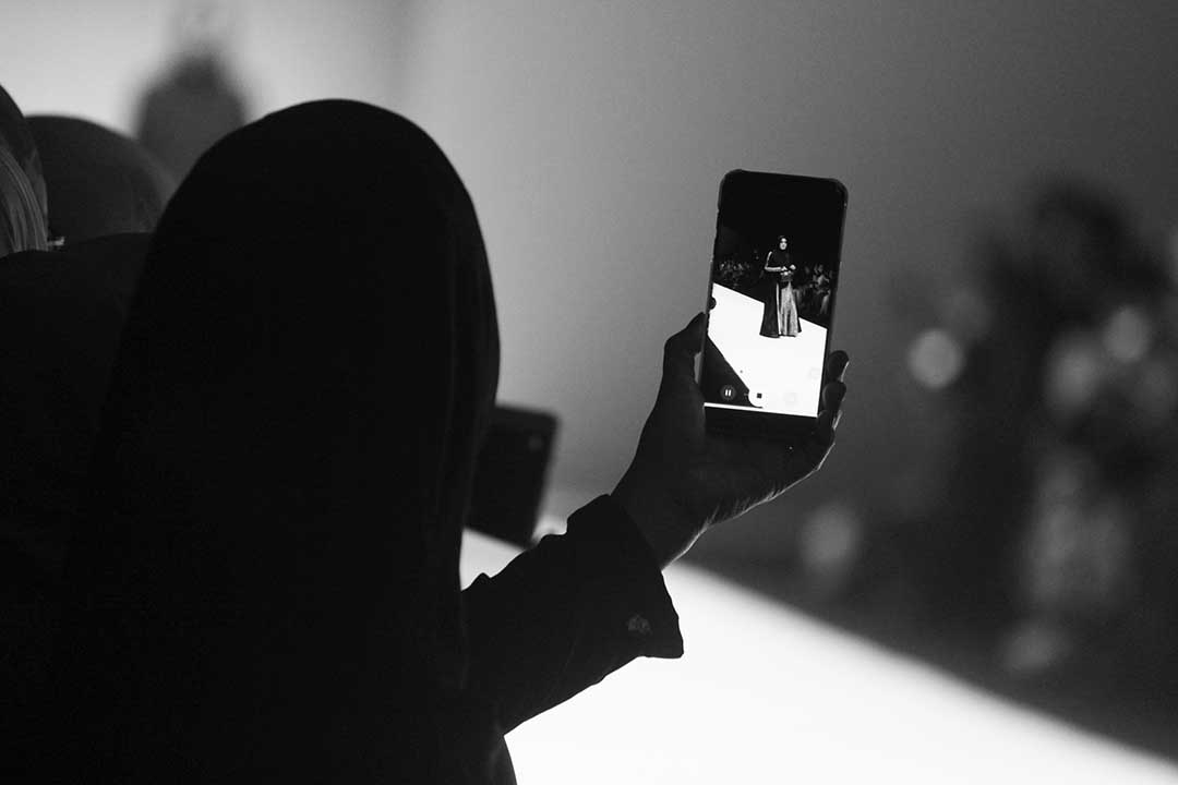 black and white image of someone holding up their phone taking a photo of a model on a runway 