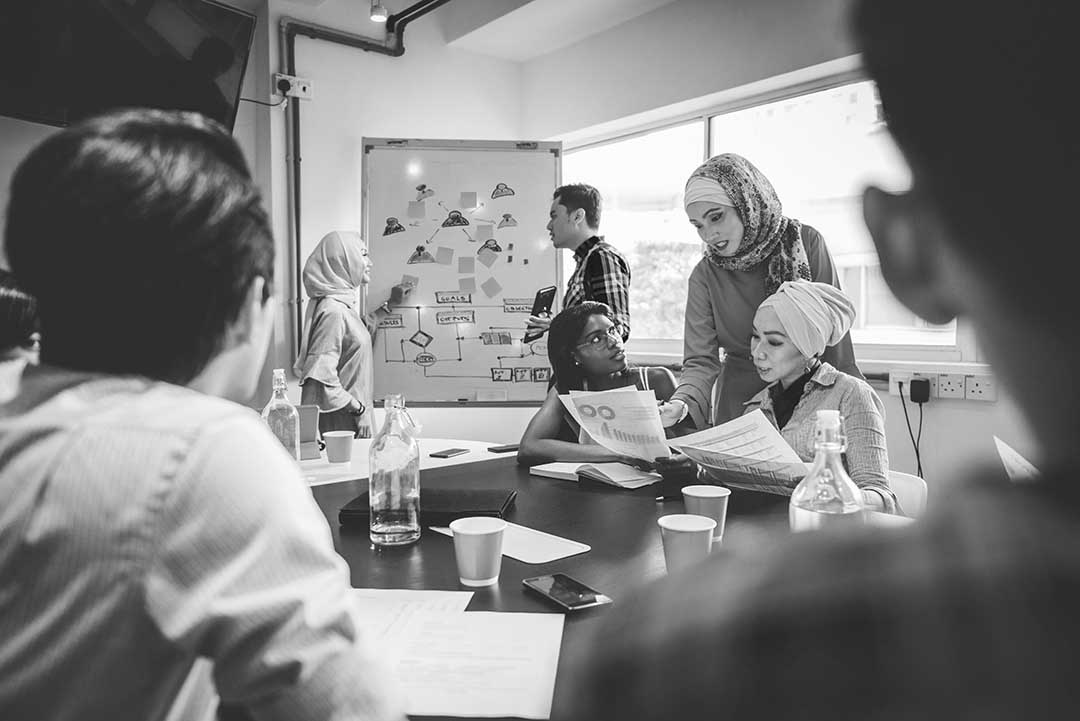 black and white image of group of people collaborating together around a table with paper and drawings on a whiteboard