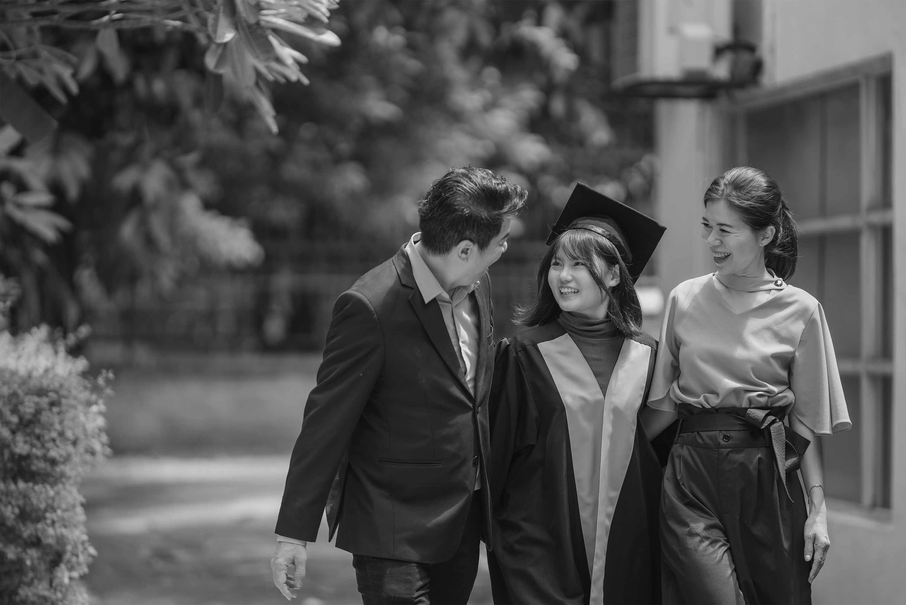 black and white image of a mother and father walking their daughter in graduation gown