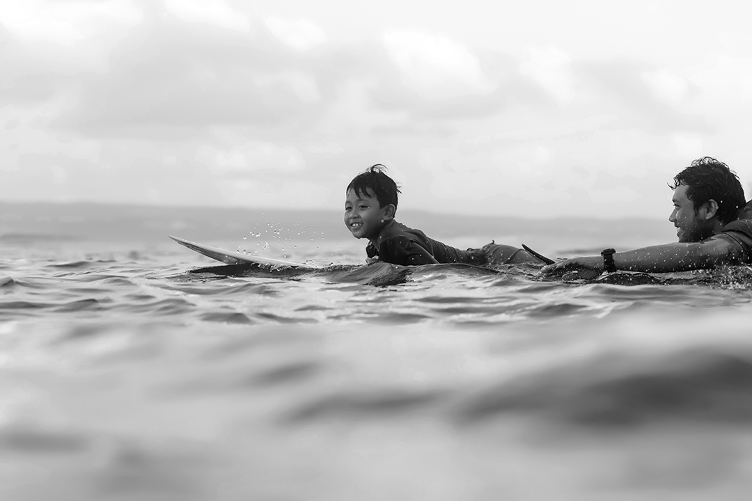 black and white image of samroze lying down smiling on a surfboard with his father pushing him