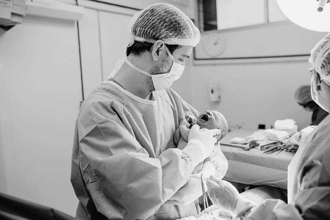 black and white photo of man in scrubs holding newborn crying in hospital 