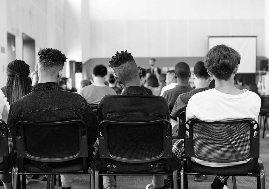 black and white image of backs of students sitting on black chairs in a classroom setting