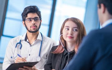 Kylie is sitting outside on a green park bench, leaning over the back of it to smile at the camera. She has long blonde hair and glasses, and is wearing a grey tweed coat.