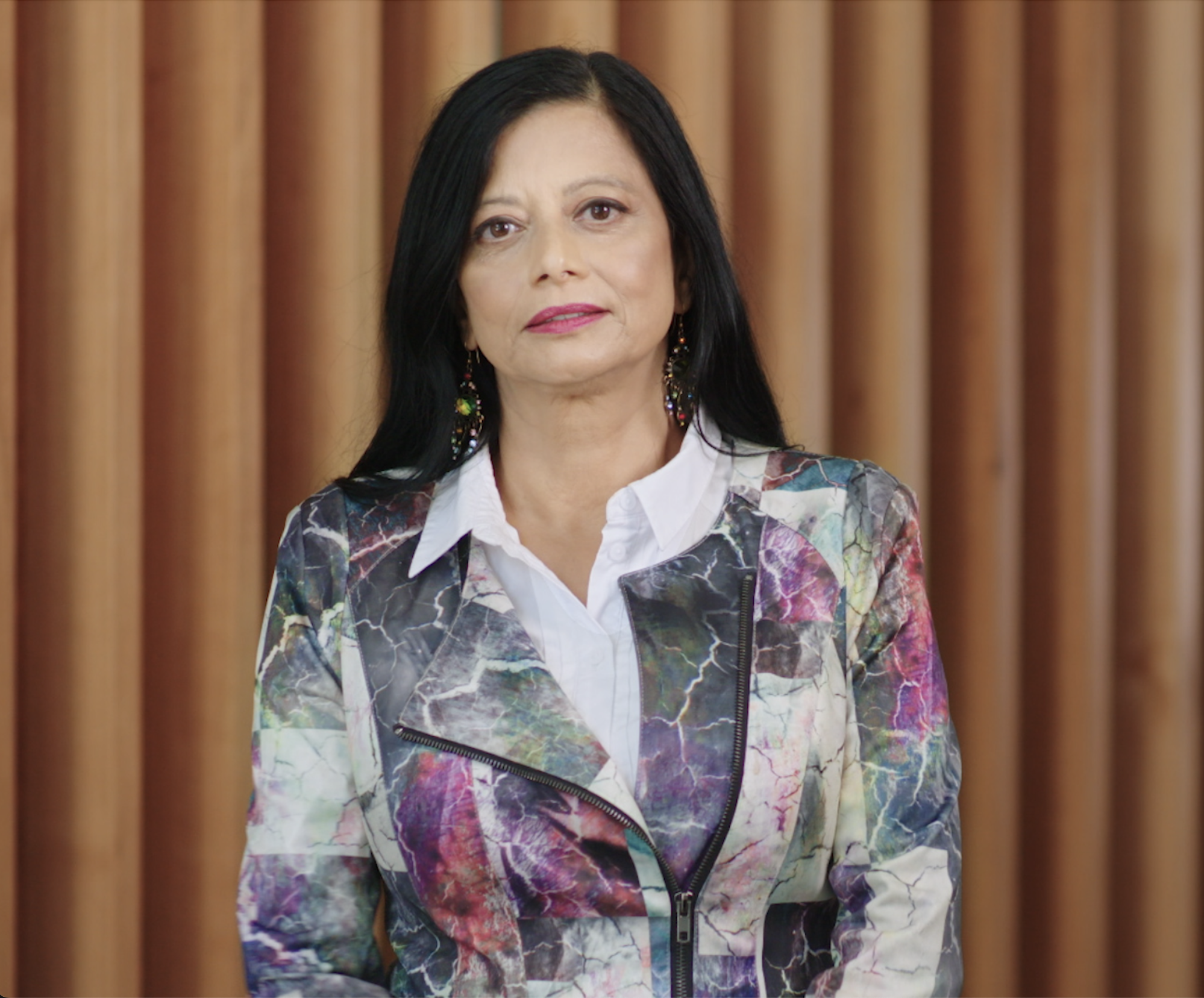 Jayashri has long black hair and is staring into the camera. She wears pink lipstick, dangling earrings, a white shirt and a black, white, purple and green floral jacket. She is standing in front of a slatted wooden background.