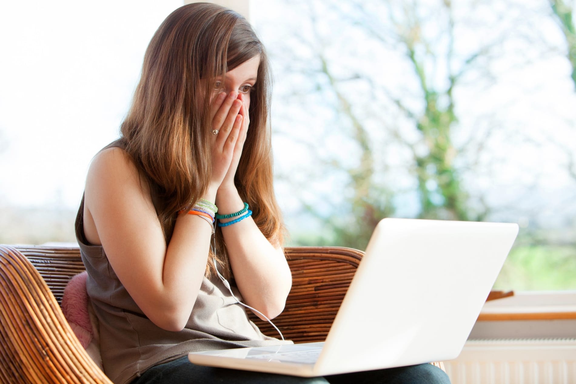 A young brown-haired woman wearing a brown top and colourful bracelets on her bare arms covers her mouth and nose with both hands as she looks at something on the white laptop in her lap. She is seated in a wicker chair and there is a window behind her.