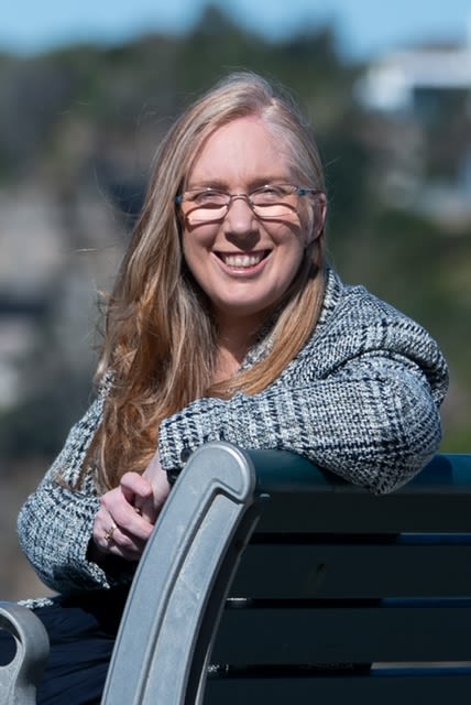 Kylie is sitting outside on a green park bench, leaning over the back of it to smile at the camera. She has long blonde hair and glasses, and is wearing a grey tweed coat.