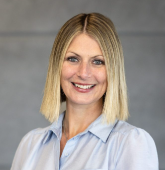 Asher Flynn has straight blonde, shoulder-length hair and is wearing a light blue collared shirt. She is smiling in front of a plain grey, industrial background.