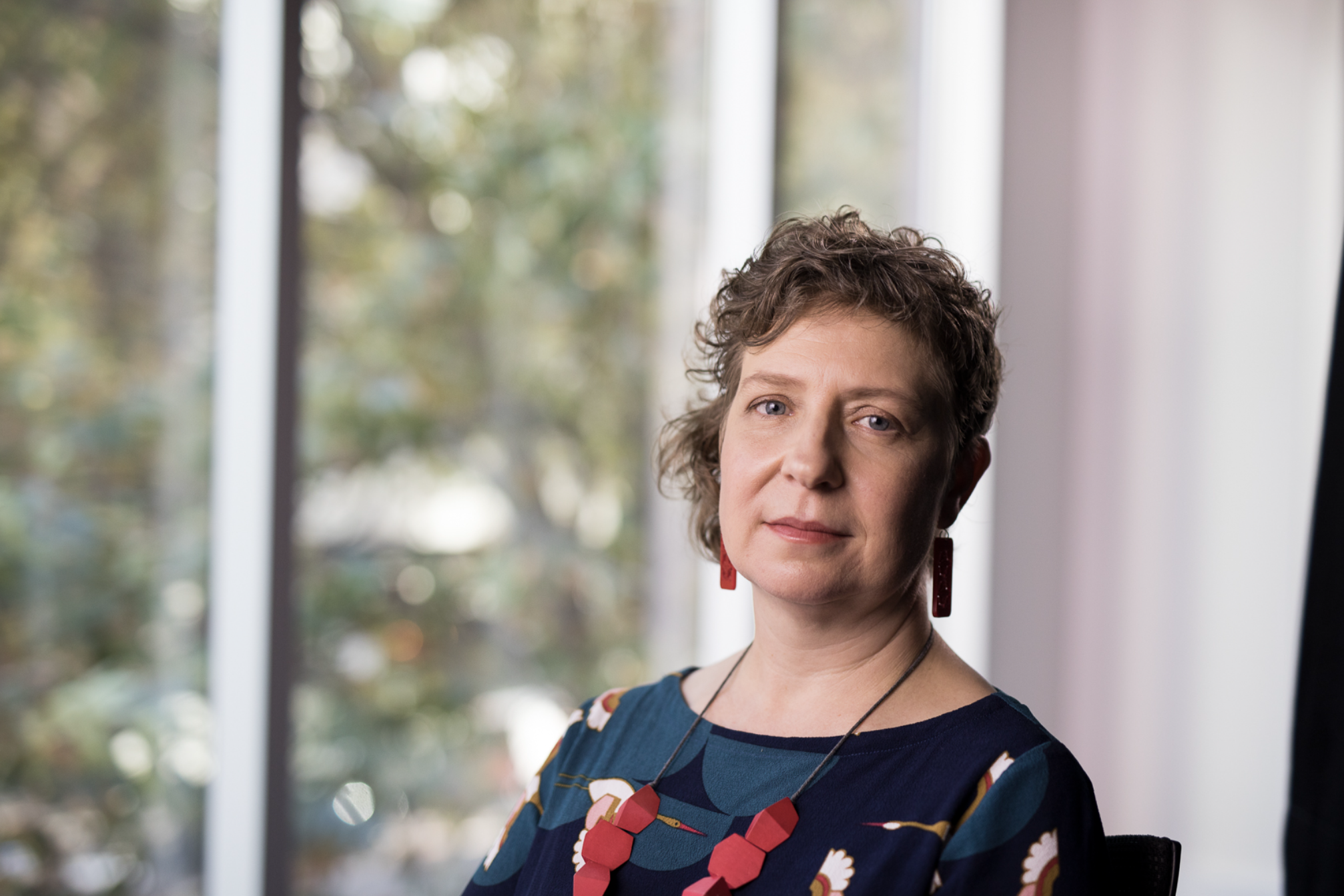 Becky has a curly, asymmetrical haircut. She is wearing dangling red earings, a necklace with large red beads, and a blue and white floral shirt. She is seated in front of a window and staring directly into the camera.