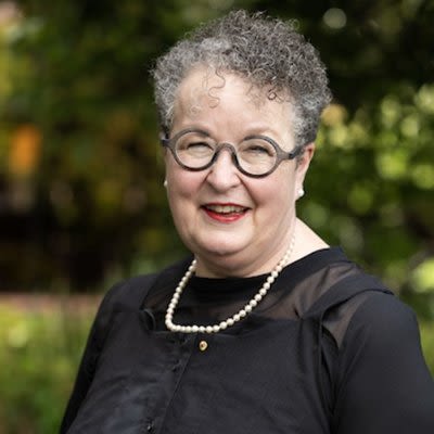 Professor Jane Fisher has short, curly grey hair. She's wearing round glasses with thick grey frames, red lipstick and a pearl necklace over a black blouse. She's smiling, and there are trees and grass in the background.