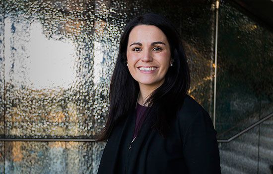Kate Fitz-Gibbon, a white woman with dark hair, smiles and poses in front of a metal wall.