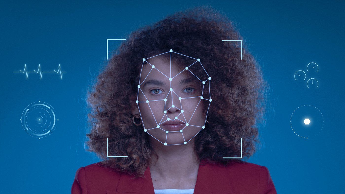 Photo-illustration of facial recognition technology analysing a woman's face. She has curly red hair and is wearing gold earrings, a red blazer and a white shirt. There are white graphics of dials and nodes overlaying her face and surrounding her against a blue background.
